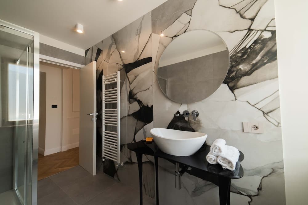 A modern bathroom features a striking black-and-white marble wall, a sleek white vessel sink on a black stand, neatly folded towels, and a round mirror, creating an elegant and sophisticated atmosphere ideal for luxury accommodation searches.