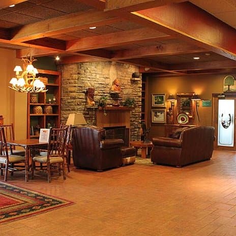 A cozy hotel lobby features warm wooden beams, a stone fireplace adorned with sculptures, leather armchairs, and a dining area, creating an inviting atmosphere ideal for travelers seeking relaxation and comfort.