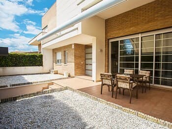 A serene suburban patio features a modern house with brick walls, glass doors, outdoor furniture, and a gravel garden, ideal for a relaxing getaway or family vacation.