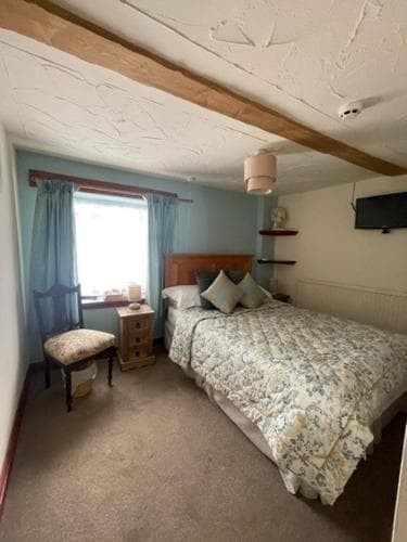 A cozy bedroom with wooden beams, a floral-patterned bedspread, and soft blue curtains creates a serene retreat, ideal for travelers seeking relaxation in a quaint lodge setting.