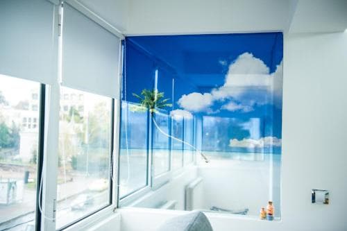 A serene hotel balcony features a vibrant blue sky-printed roller blind with fluffy clouds, enhancing the modern urban view through large glass windows, ideal for travelers seeking a calming retreat in a bustling city.