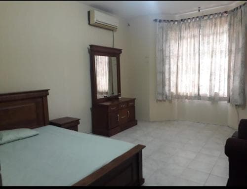 A cozy bedroom with tiled flooring, wooden furniture including a bed and dresser with a mirror, sheer curtains covering a window, and an air conditioner, ideal for travelers seeking a simple and functional accommodation.