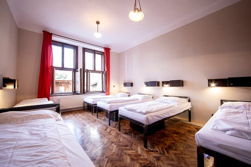 A cozy hostel room in Prague features multiple beds with white linens, warm wooden flooring, red curtains framing a bright window, and ambient lighting, ideal for budget travelers seeking comfort and simplicity.