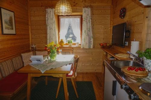 A cozy wooden cabin interior features a rustic dining area with vibrant flowers on the table, a kitchenette with fresh fruit and pastries, and a window view of a snow-capped mountain, ideal for travelers seeking a serene retreat in nature.