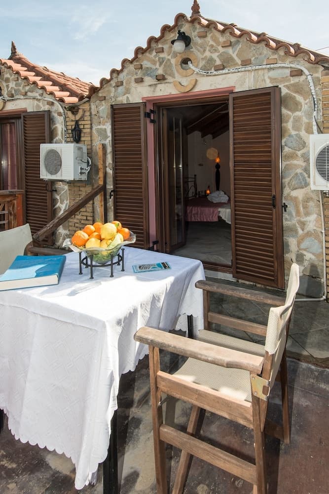 A cozy stone cottage patio with wooden shutters, a fruit-filled tablecloth-covered table, and inviting chairs creates a serene vacation retreat perfect for relaxation and leisurely dining.