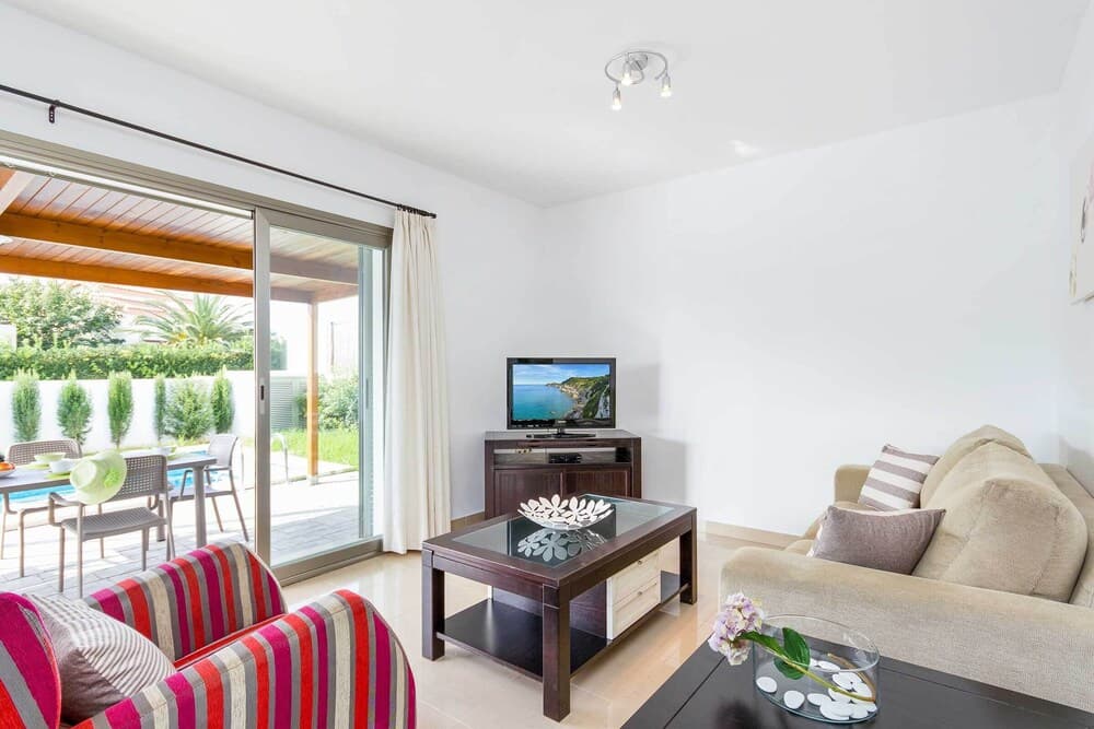 A bright, modern living room with a cozy sofa, colorful armchair, and a glass coffee table overlooks a serene outdoor patio with lush greenery, ideal for travelers seeking a relaxing vacation rental experience.