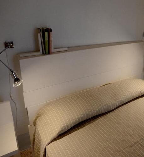 A cozy bedroom scene featuring a neatly made bed with striped bedding, a minimalist white headboard, a few books, and a modern wall-mounted lamp, ideal for travelers seeking a tranquil and stylish accommodation option.