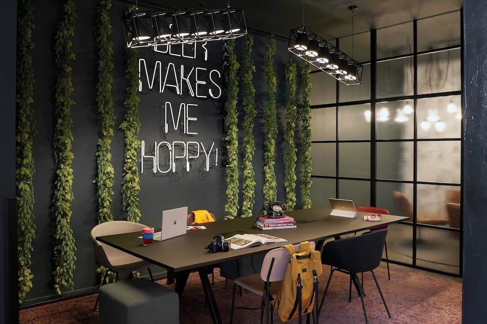A cozy co-working space features a vibrant neon sign reading "Beer Makes Me Hoppy" amidst lush greenery, modern lighting, and a communal table set with laptops and books, ideal for travelers seeking a creative and inspiring work environment.