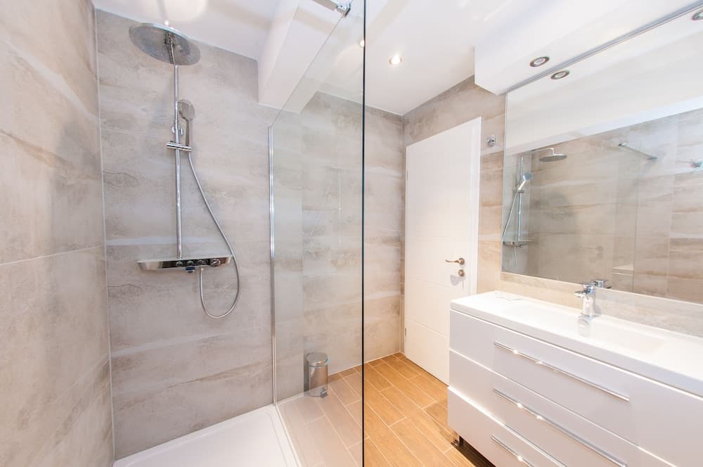 A modern bathroom with sleek beige tiles, a glass-enclosed shower, white vanity, and large mirror exudes a clean and serene atmosphere, ideal for travelers seeking a luxurious accommodation experience.