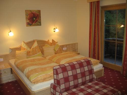 A cozy hotel room features a warmly lit bed with orange bedding, a checkered bench, and a view of greenery through a glass door, ideal for travelers seeking comfort and relaxation.