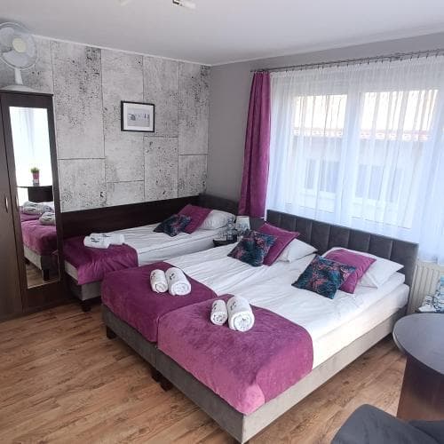 A cozy hotel room features two neatly made beds adorned with purple linens, a textured gray wall, and large windows draped with sheer curtains, creating a welcoming atmosphere ideal for travelers seeking comfort and relaxation.