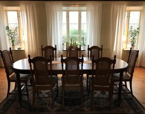 A cozy dining room with wooden tables and chairs set on a patterned rug, framed by large windows with sheer curtains, evokes a serene and inviting atmosphere ideal for family gatherings or intimate dinners.