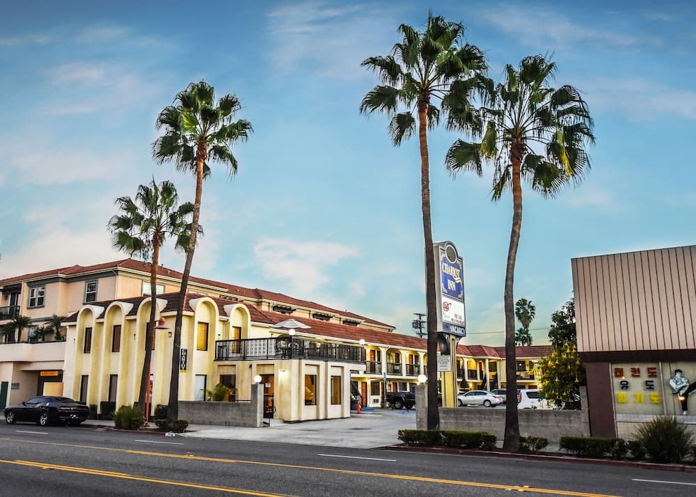 A sunny roadside scene featuring a charming motel with palm trees, a "Chariot Inn" sign, and nearby Korean signage, ideal for travelers seeking a cozy stay in a vibrant urban area.