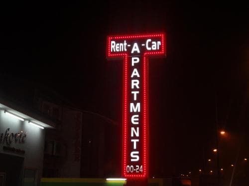 A vibrant neon sign reading "Rent-A-Car APARTMENTS" illuminates the night sky above a building, creating an inviting urban atmosphere ideal for travelers seeking accommodation or car rental services.