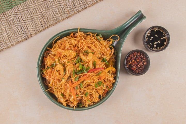 A vibrant bowl of noodles garnished with vegetables and shrimp sits invitingly on a beige tablecloth at Jugnoo Cafe in Gurugram, India, accompanied by small bowls of chili flakes and sesame seeds, perfect for food enthusiasts exploring local dining options.
