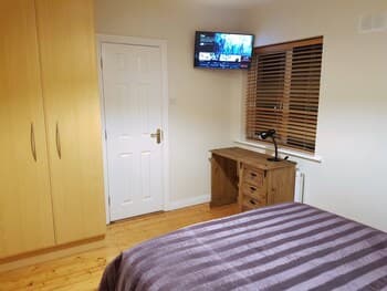 A cozy bedroom with wooden flooring, a neatly made bed, a desk by the window with blinds, and a mounted TV creates a serene atmosphere ideal for travelers seeking comfort and relaxation.