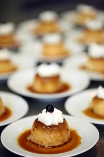 Rows of caramel-flavored desserts topped with whipped cream and blueberries on white plates create an inviting atmosphere in a bustling bakery, perfect for travelers seeking a sweet culinary experience.