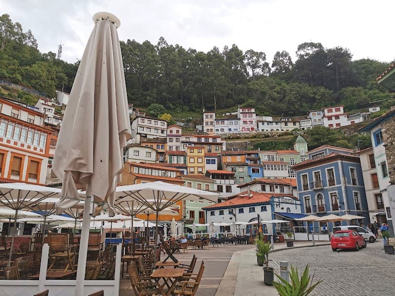 A charming village square in Asturias, Spain, features colorful houses on a hillside, outdoor seating under umbrellas, and a tranquil atmosphere ideal for leisurely strolls and cultural exploration.