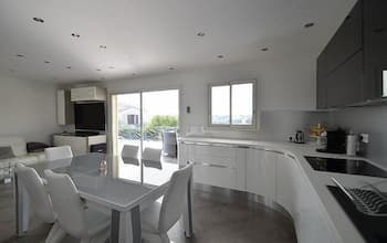 A modern, minimalist kitchen and dining area with sleek white furniture, large windows letting in natural light, and a cozy yet stylish atmosphere ideal for a family vacation rental.