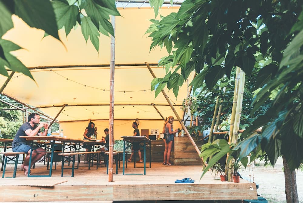 A serene outdoor dining area under a bamboo-framed canopy with lush greenery, featuring wooden tables where people enjoy meals, ideal for eco-friendly retreats and nature lovers.