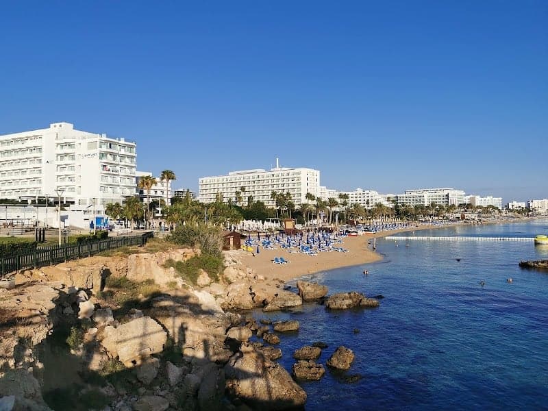 A serene coastal scene at a beach hotel in Cyprus features clear blue skies, white buildings, palm trees, colorful beach umbrellas, and calm turquoise waters, ideal for a relaxing vacation getaway.