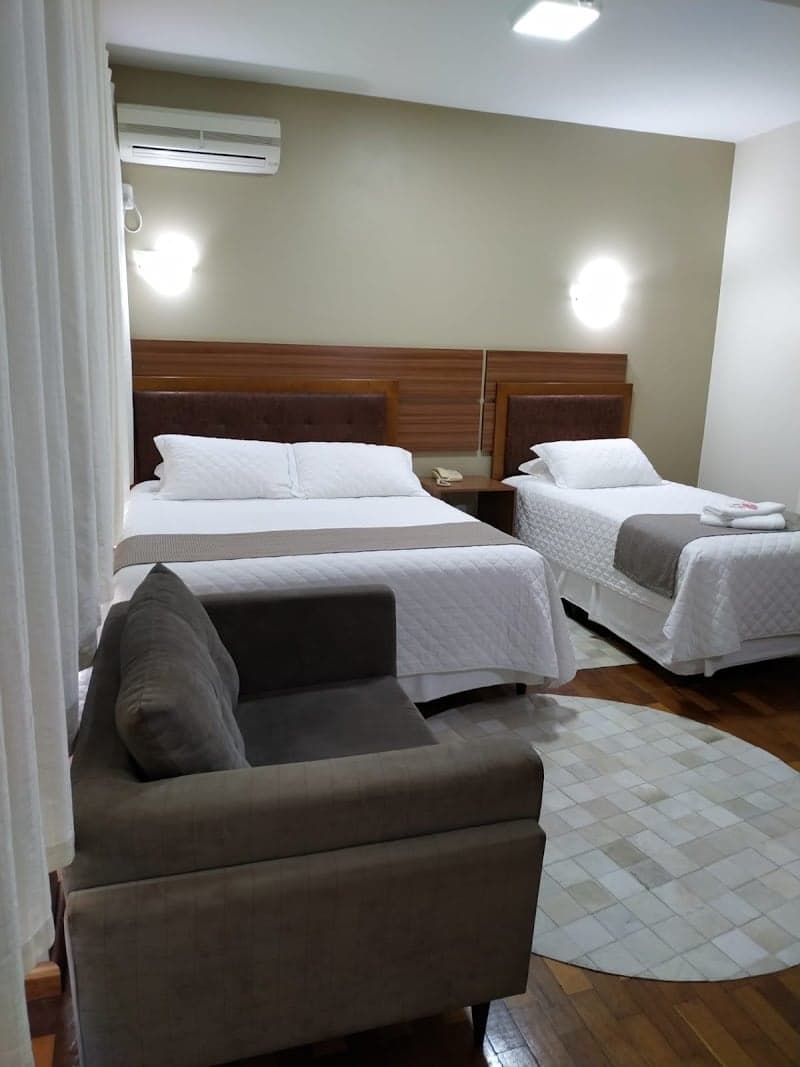 A cozy hotel room at Maravilha Palace Hotel in Brazil features two neatly made beds with wooden headboards, a comfortable armchair, soft lighting, and a warm ambiance ideal for travelers seeking relaxation.
