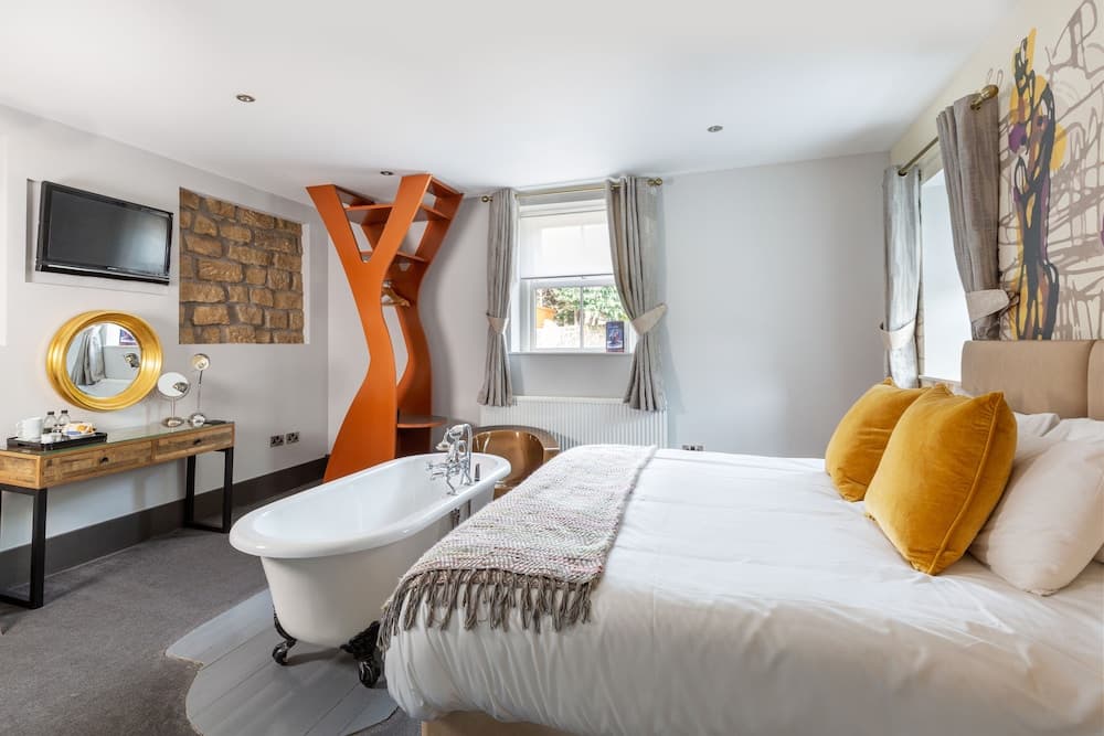 A cozy hotel room features a stylish freestanding bathtub, vibrant orange spiral staircase, and a plush bed adorned with mustard yellow pillows, ideal for travelers seeking comfort and unique design elements.