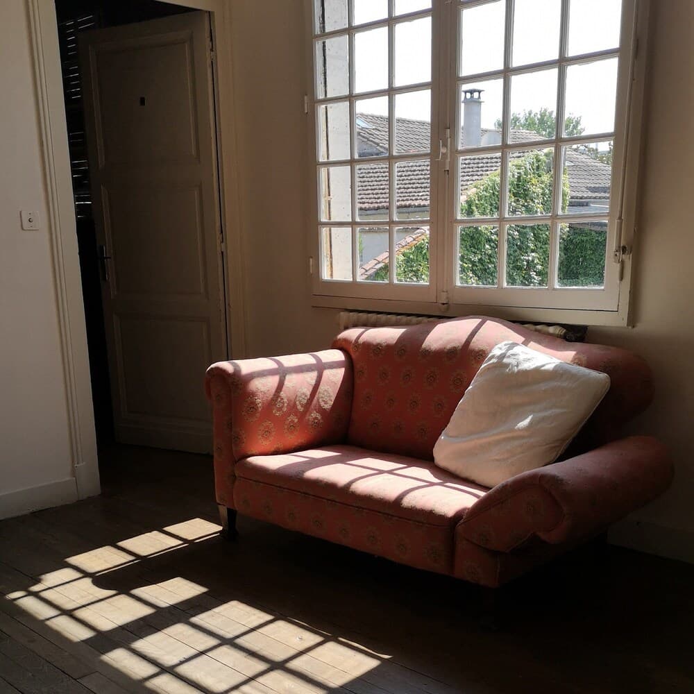 A cozy vintage sofa bathed in sunlight through a large window offers a serene retreat in a rustic European cottage, perfect for travelers seeking a peaceful getaway.