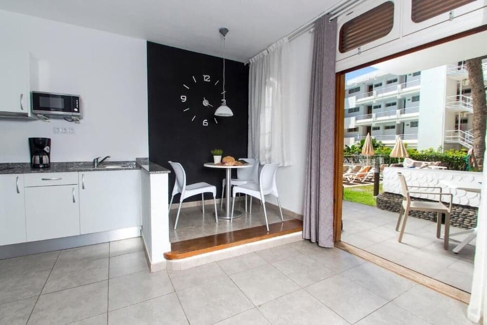 A cozy apartment interior features a modern kitchenette, a dining area with a bold black wall clock, and an inviting balcony view of a sunny resort patio, perfect for travelers seeking comfort and convenience in a beachside retreat.