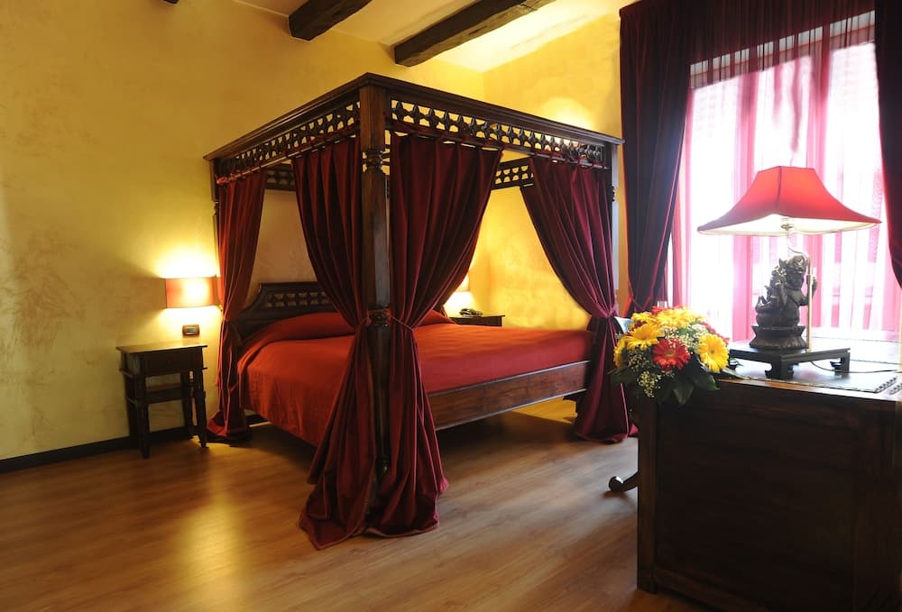 A cozy hotel room features a majestic four-poster bed draped in rich red curtains, warm yellow walls, wooden floors, and vibrant floral arrangements, creating an inviting atmosphere perfect for travelers seeking comfort and elegance.