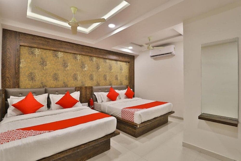 A cozy hotel room features two neatly arranged beds with red accents against a tree-patterned wall, complemented by modern ceiling fans and an air conditioner, ideal for travelers seeking comfort and convenience.