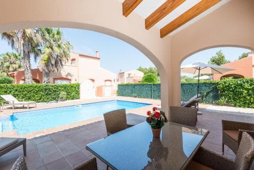 A serene Mediterranean villa patio features a sparkling pool, lush greenery, and cozy seating under a wooden-beamed archway, ideal for a relaxing vacation retreat.