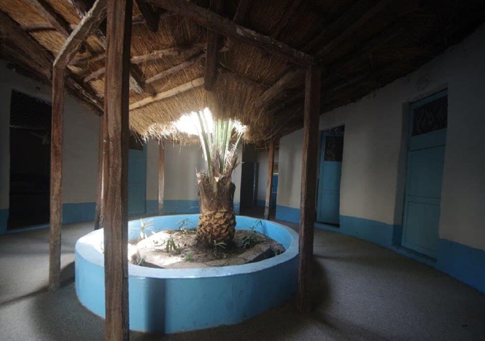 A serene courtyard features a central palm tree in a blue planter under a thatched roof, surrounded by rustic wooden pillars and blue-trimmed doors, ideal for cultural heritage tourism.