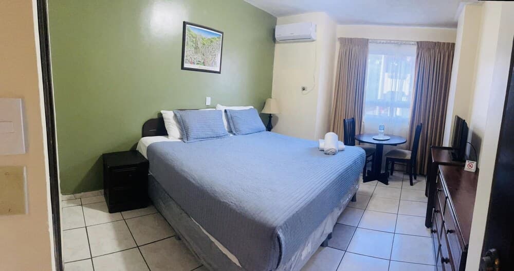 A cozy hotel room with a neatly made bed, green walls, and a small dining area by the window offers a comfortable stay for travelers seeking relaxation.
