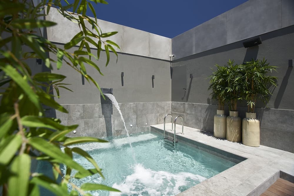 A serene courtyard pool features a cascading water feature against minimalist concrete walls, surrounded by lush greenery in large pots, ideal for a tranquil urban retreat experience.