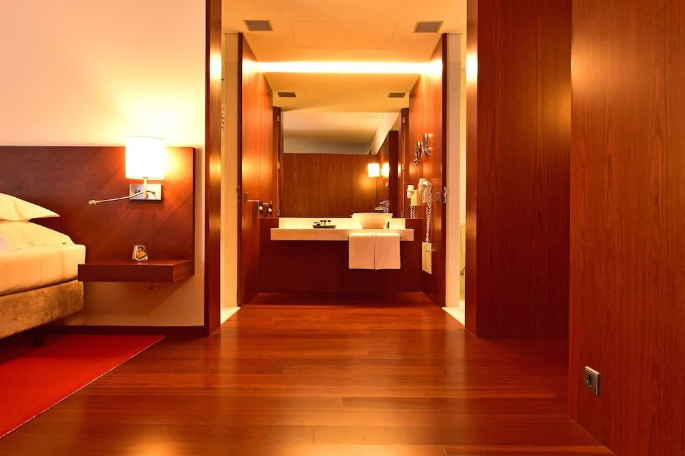 A cozy hotel room with warm wooden tones, a neatly made bed, and an inviting bathroom featuring a double sink and soft lighting, perfect for travelers seeking comfort and relaxation.