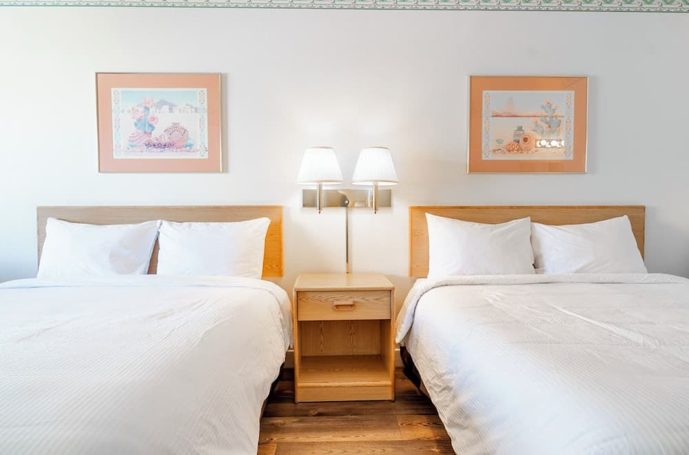 A cozy hotel room features two neatly made beds with white linens, framed artwork on the walls, and a wooden nightstand with lamps, ideal for travelers seeking comfort and simplicity.