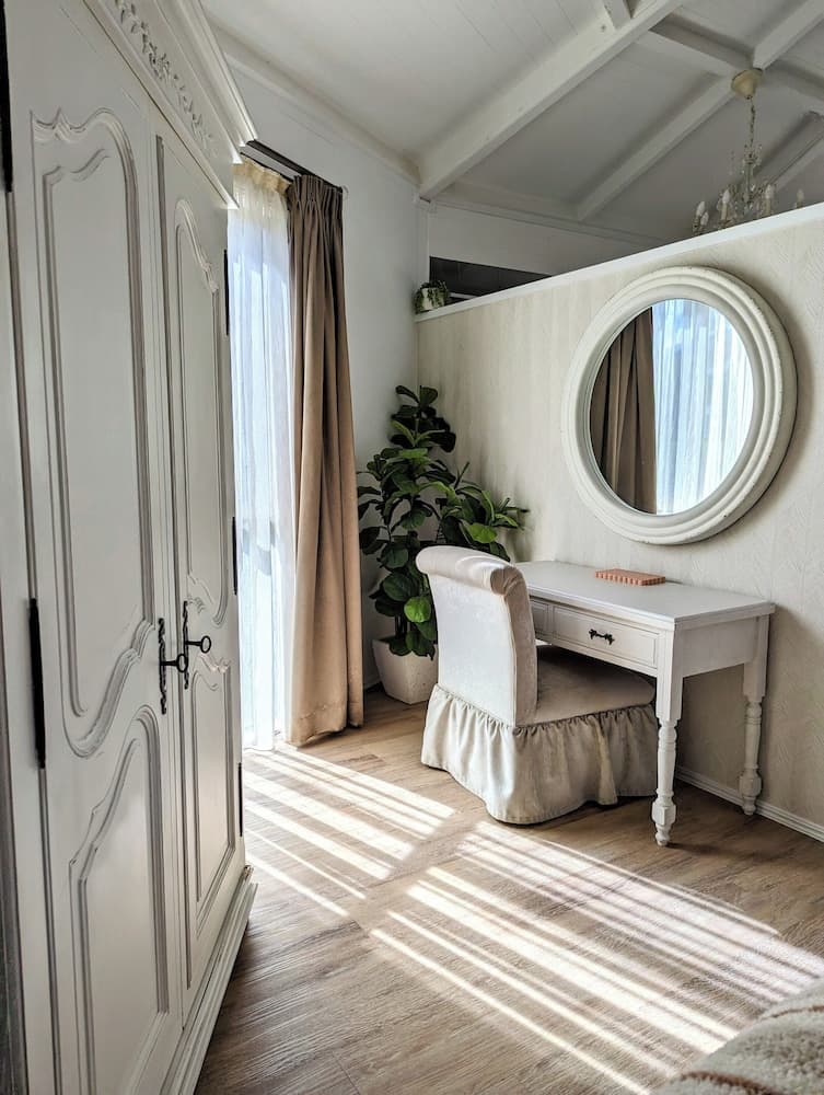 A serene, sunlit bedroom corner features a vintage white vanity with a round mirror, a cozy upholstered chair, lush greenery, and elegant cabinetry, ideal for travelers seeking a charming and relaxing retreat.