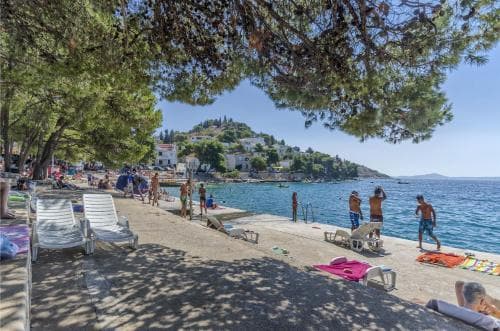 A serene coastal beach scene with sunbathers relaxing on lounge chairs under the shade of lush pine trees, set against a backdrop of turquoise waters and distant hills, ideal for travelers seeking a tranquil seaside getaway.