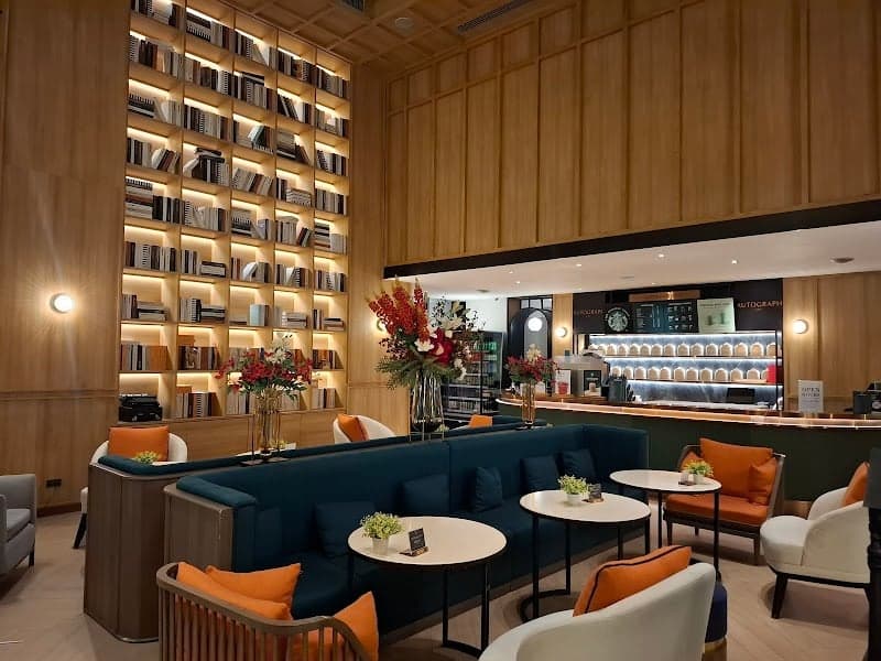 A cozy hotel lounge with wooden walls, bookshelves, plush seating, and vibrant decor offers a serene retreat for travelers seeking relaxation in Bangkok's urban setting.