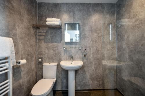 A modern bathroom with sleek gray walls, a pedestal sink, glass shower enclosure, and neatly arranged towels exudes a clean and sophisticated ambiance, ideal for travelers seeking comfort in upscale accommodations.