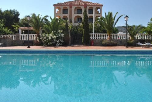 A serene resort scene features a tranquil pool reflecting a multi-story villa surrounded by lush palm trees and blooming shrubs, ideal for a relaxing vacation getaway.