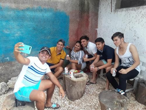 A cheerful group of friends gathers outdoors on a rustic patio, enjoying drinks and taking selfies against a colorful wall backdrop, perfect for capturing authentic travel moments and social bonding experiences.