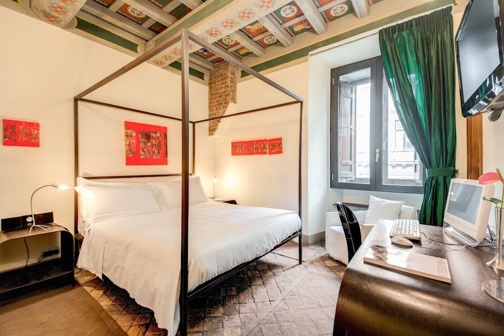 A cozy hotel room with a canopy bed, vibrant red artwork, a patterned ceiling, and a desk setup by the window, ideal for travelers seeking comfort and a touch of elegance.