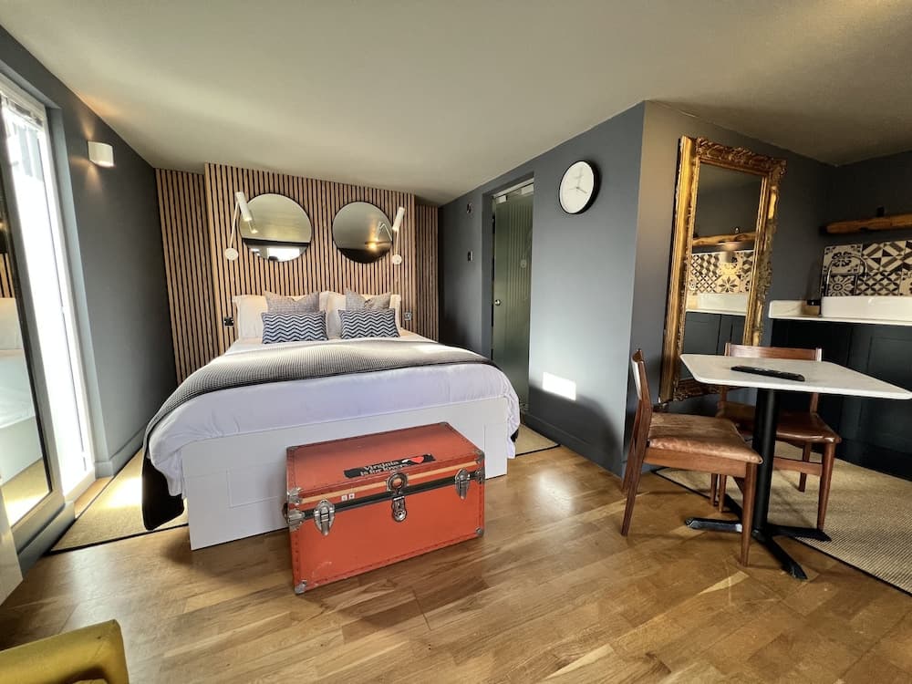 A cozy hotel room features a stylish bed with wooden slat headboard, vintage orange trunk, elegant gold-framed mirror, and a small dining area, creating a warm and inviting atmosphere ideal for travelers seeking comfort and charm.