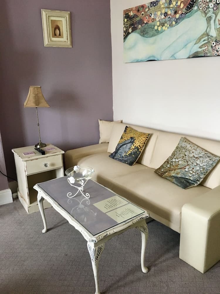 A cozy living room corner features a beige sofa adorned with patterned cushions, a vintage-style coffee table with magazines, a small side table with a lamp, and artistic wall decor, creating an inviting atmosphere ideal for relaxation or a quiet getaway.