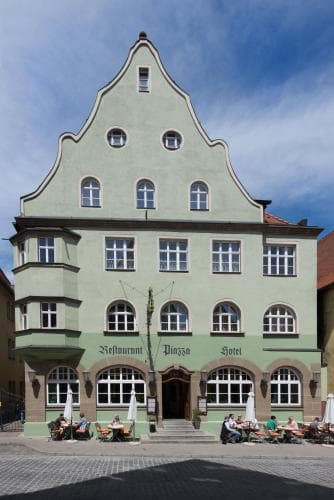 A charming European town square features a historic green building with arched windows and outdoor seating, ideal for travelers seeking a cozy café or hotel experience in a picturesque setting.