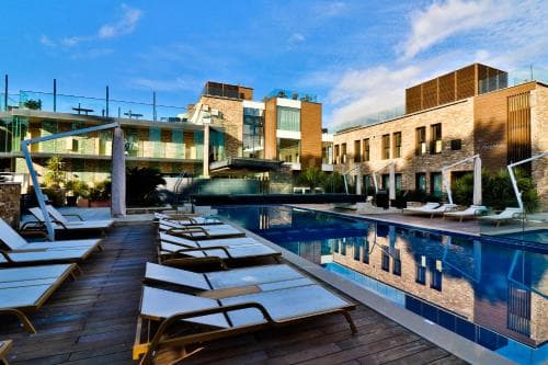 A serene rooftop pool area with modern lounge chairs and reflective water, surrounded by contemporary buildings under a clear blue sky, ideal for luxury hotel relaxation and vacation photography.
