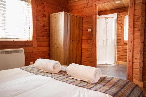 A cozy cabin bedroom with wooden walls, a neatly made bed adorned with plaid bedding and rolled towels, a compact shower enclosure, and natural light streaming through the window, ideal for travelers seeking a rustic retreat.