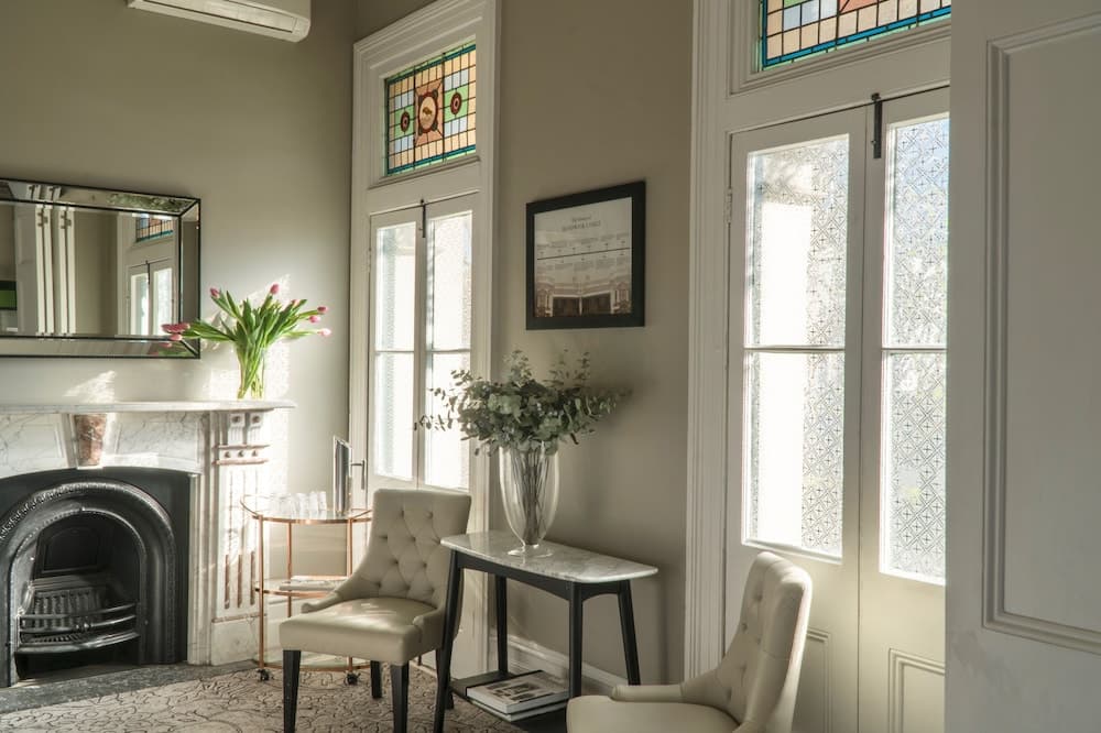 A cozy, sunlit room with elegant tufted chairs, stained glass windows, and a marble fireplace creates an inviting atmosphere perfect for a relaxing retreat or boutique hotel stay.